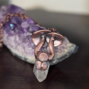 May include: A copper-colored mushroom pendant necklace with a chain, set against a backdrop of a purple amethyst crystal. The pendant features two detailed mushroom caps and stems, attached to a clear crystal point.