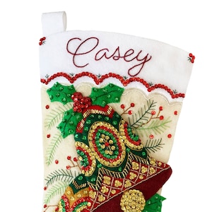 May include: A white felt Christmas stocking with the name "Casey" embroidered on the top. The stocking is decorated with three red, green, and gold bells with sequins and beads. The bells are surrounded by green holly leaves and red berries.