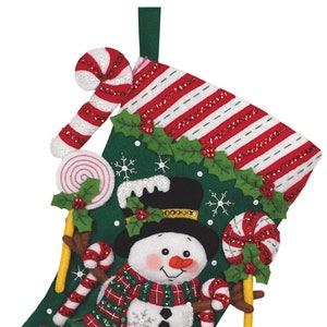 Könnte beinhalten: Ein Weihnachtsstrumpf mit einem Schneemann mit schwarzem Zylinder, rotem Schal und Zuckerstangenarmen. Der grüne Strumpf ist mit Süßigkeiten, Stechpalmen und Schneeflocken verziert.