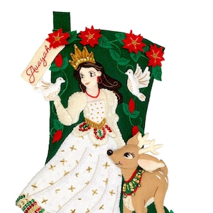 Op de afbeelding: Een kerstsok met een prinses met een kroon, die duiven en een rendier vasthoudt. De sok is groen met rode poinsettia's en de naam "Avayah". De prinses draagt een witte jurk met gouden accenten.