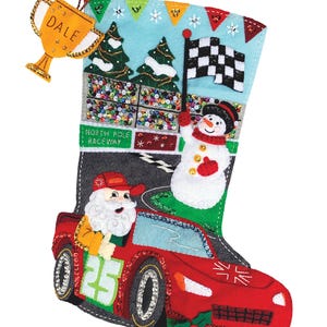Puede incluir: Una media de Navidad de fieltro con un coche de carreras rojo, Papá Noel conduciendo y un muñeco de nieve sosteniendo una bandera a cuadros. La media tiene el texto "North Pole Raceway" y "Arctic Rush". Un trofeo dorado con el nombre "Dale" está unido a la parte superior de la media.