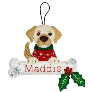 Può includere: Addobbo natalizio in feltro con un labrador retriever giallo che indossa un maglione rosso e verde, seduto su un osso bianco con il nome "Maddie" ricamato su di esso. L'osso è decorato con fiocchi di neve e un ramo di agrifoglio con bacche rosse.