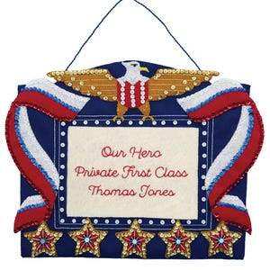 Puede incluir: Adorno de pared patriótico con fondo azul y marco blanco. Presenta un águila dorada, cintas rojas y blancas, y cinco adornos en forma de estrella. El texto dice "Our Hero Private First Class Thomas Jones."