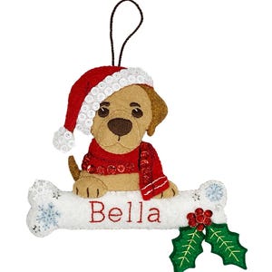 Può includere: Addobbo natalizio in feltro di un cane marrone che indossa un cappello da Babbo Natale rosso e una sciarpa, seduto su un osso bianco con il nome "Bella" ricamato su di esso. L'osso è decorato con fiocchi di neve e agrifoglio.