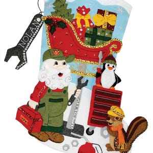 Peut inclure: Une chaussette de Noël en feutre avec un fond bleu et un personnage de Père Noël tenant une clé à molette. La chaussette présente un traîneau rouge avec des cadeaux, un pingouin et un écureuil. La chaussette porte une étiquette qui dit "NOLAN".