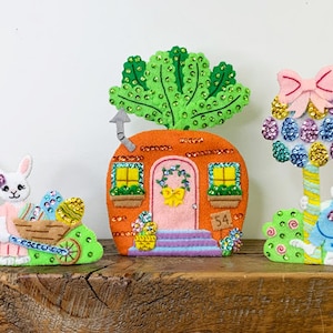 May include: Six colourful Easter-themed figurines featuring bunnies, a carrot house, an egg, and a flower-filled car. The figurines are made of felt and decorated with rhinestones.