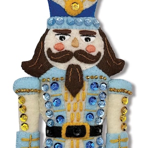 May include: A felt Christmas ornament shaped like a nutcracker soldier. The ornament features a blue and white uniform with gold and blue embellishments, a brown mustache, and a blue crown. A gold string is attached for hanging.