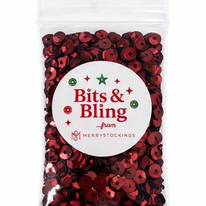Op de afbeelding: Een doorzichtige plastic zak gevuld met glanzende, rode pailletten. Een wit rond etiket met de tekst "Bits & Bling" in het rood, met groene en rode accenten, en de tekst "...from MerryStockings".