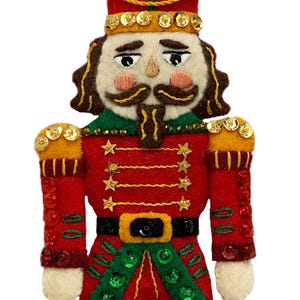 May include: A felt Christmas ornament depicting a nutcracker soldier. The ornament is red, green, and gold with a white face and black boots. The nutcracker is wearing a red and gold uniform with a black belt and green trim. The ornament has a gold string for hanging.