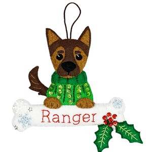 Può includere: Addobbo natalizio in feltro di un cane marrone che indossa un maglione verde con paillettes, seduto su un osso bianco con il nome "Ranger" ricamato su di esso. L'osso è decorato con fiocchi di neve e un ramo di agrifoglio con bacche rosse.