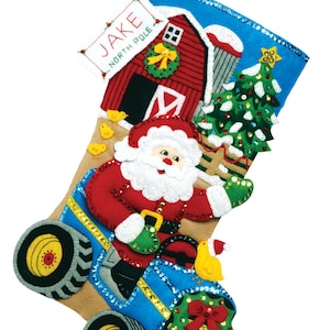 May include: A felt Christmas stocking with a blue background and a scene of Santa Claus driving a blue tractor with a red barn and a Christmas tree in the background. The stocking is personalized with the name "JAKE NORTH POLE".
