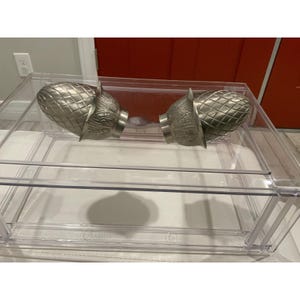 May include: Two silver decorative finials with a pineapple design. They are displayed inside a clear, rectangular acrylic box. The finials have a textured surface and are designed to be attached to a curtain rod.