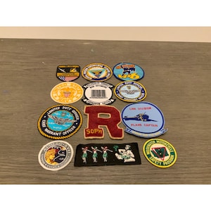 Puede incluir: Una colección de doce parches de tela bordados con diversos emblemas militares, de aviación y organizativos. Los parches presentan formas circulares, rectangulares y de escudo con una variedad de colores, textos y diseños gráficos.
