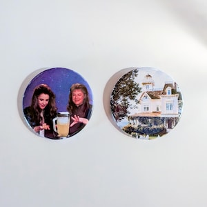 May include: Two circular pin-back buttons. The left button features two individuals with dark hair standing before a purple background, one holding a glass pitcher. The right button displays a detailed illustration of a large white Victorian house.