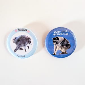 May include: Two circular pin-back buttons. The left button has a light blue border, a raccoon image, and the text JIMOTHY FAN CLUB with two small pink hearts. The right button has a dark blue background, a raccoon image, and the text WEIRD LITTLE CREATURE CLUB.