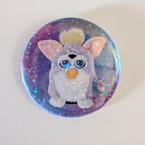 May include: A round button featuring a Furby character with blue eyes, a yellow beak, and pink ears. The background is a gradient of purple and blue with star-shaped sparkles. A small image of a Furby is in the lower right corner.
