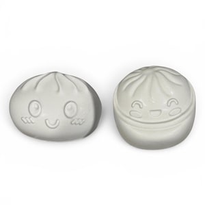 May include: Two off-white ceramic dumpling-shaped containers with smiling faces. One is a full dumpling, the other is a dumpling with a lid. The containers have a dimpled texture and are designed to resemble traditional Asian dumplings.