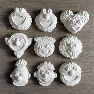 May include: A collection of nine white, unpainted plaster figurines shaped like various Muppet character faces. The set includes recognizable figures such as Cookie Monster, Elmo, Miss Piggy, Count von Count, Oscar the Grouch, Big Bird, Bert, Ernie, and Fozzie Bear, all featuring detailed molded facial expressions.