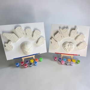 May include: Two white canvases on wooden easels featuring raised hand sign sculptures. One canvas displays a superhero mask, the other a heart with a bow. Each set includes a paintbrush and a strip of small paint pots in various colors.