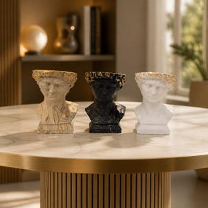 Puede incluir: Tres bustos decorativos de figuras clásicas. Uno es blanquecino con detalles dorados, el segundo es negro y el tercero es blanco. Todos tienen una corona dorada. Se exhiben sobre una mesa redonda con tapa de mármol y borde dorado.