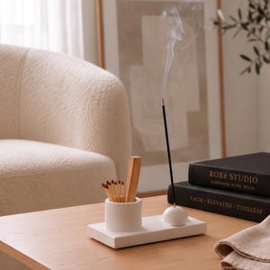 May include: A white rectangular incense holder tray featuring a cylindrical matchstick container and a small dome-shaped incense stick holder. A thin black incense stick is inserted into the dome, emitting a thin trail of white smoke.