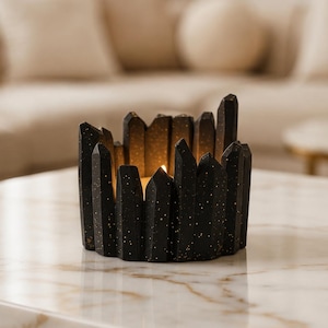 May include: A black candle holder with a crystal design. The holder has gold speckles and is on a marble table. A lit candle inside provides a warm light. Home decor item.