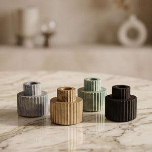 Puede incluir: Cuatro candelabros decorativos en tonos grises, beige, verdes y negros. Cada soporte tiene una base cilíndrica con ranuras verticales y una parte superior cilíndrica más pequeña. Los candelabros están dispuestos sobre una superficie de mármol.