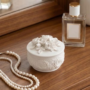 May include: A round white ceramic trinket box featuring a lid adorned with sculpted floral rose details and a base decorated with a relief pattern of roses and leaves. The box is positioned near a string of white pearls and a glass perfume bottle.