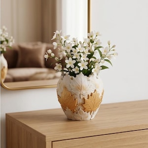 Può includere: Un vaso decorativo color crema con foglie dorate, pieno di fiori bianchi. I fiori hanno piccoli petali bianchi e steli verdi. Il vaso è appoggiato su una superficie di legno.
