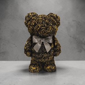 Könnte beinhalten: Goldene Teddybär-Figur mit aufwendigem Rosenmuster. Der Bär hat eine silberne Fliege und steht auf einer weißen Oberfläche vor grauem Hintergrund. Die Gesamtästhetik ist elegant und dekorativ.