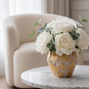 Puede incluir: Un arreglo floral con grandes peonías blancas y follaje verde en un jarrón dorado con diseño de hojas. El jarrón está sobre una mesa con tapa de mármol, con una silla color crema en el fondo.