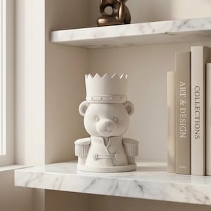 May include: A matte off-white ceramic figurine of a bear wearing a crown and a military-style jacket with epaulets. The bear has a rounded face, small ears, and a simple facial expression. The figurine is positioned on a white marble shelf.
