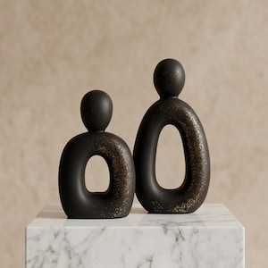 Op de afbeelding: Twee zwarte abstracte sculpturen met een matte afwerking en gouden spikkels. De sculpturen staan op een wit marmeren oppervlak. Eén is ovaal met een cirkelvormige opening, de andere is een langwerpige vorm.