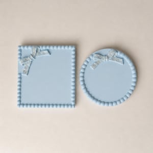 May include: Two light blue decorative trays, one square and one round. Each tray features a beaded border and a bow with a floral pattern. The square tray has a side length of approximately 15 cm, and the round tray has a diameter of approximately 13 cm.