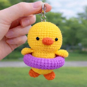 Puede incluir: Un llavero de pato amarillo hecho a mano con un pico y patas naranjas, y un flotador morado. El pato tiene ojos negros y está sujeto a un llavero plateado.