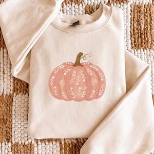 May include: A cream-colored crewneck sweatshirt featuring a central graphic of a muted pink pumpkin. The pumpkin is decorated with a delicate white line-art pattern of small flowers, leaves, and stems, and has a brown stem at the top.