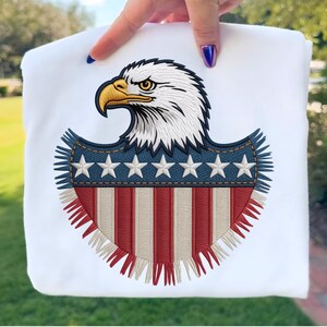 May include: White sweatshirt featuring an embroidered design of a bald eagle's head and a shield. The shield has a blue denim-like border with white stars, and red and white stripes. The eagle has white feathers, a yellow beak, and a focused gaze.