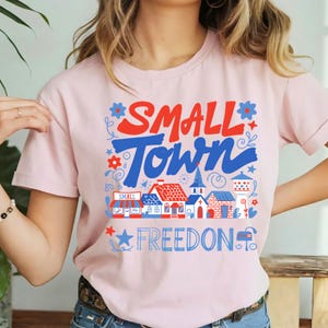 Könnte beinhalten: Hellrosa T-Shirt mit dem Schriftzug "SMALL TOWN FREEDOM" in Rot und Blau, sowie einer Illustration einer Kleinstadt. Das Design zeigt Häuser, eine Kirche und dekorative Elemente.