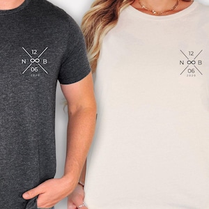 Könnte beinhalten: Ein anthrazitfarbenes T-Shirt und ein cremefarbenes T-Shirt, beide mit einer schwarzen Grafik auf der linken Brust. Die Grafik besteht aus einem Unendlichkeitssymbol zwischen den Buchstaben N und B sowie den Zahlen 12, 06 und 2020.