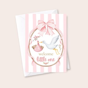 May include: A greeting card with a pink and white striped background featuring a watercolor illustration of a stork carrying a teddy bear in a pink bundle. The design includes a large pink bow, a wooden hoop frame, and the text welcome little one.