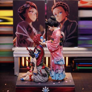 Puede incluir: Un conjunto de figuras detalladas que representan a dos personajes de anime. Uno está arrodillado, con cabello rojo y marcas azules, mientras que el otro, con un kimono rosa, le toca la cara. El fondo presenta arte de anime.