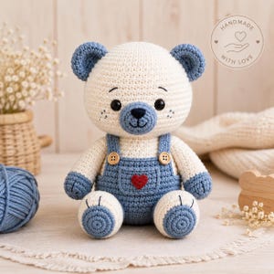 Peut inclure: Un ours en peluche crocheté avec une fourrure crème, des oreilles, des pattes et un museau bleus. L'ours porte une salopette bleue avec deux boutons en bois et un petit cœur rouge cousu sur la poche avant. Il a des yeux noirs et un nez brodé.