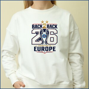 Pode incluir: Uma camisola branca de gola redonda com uma impress&atilde;o gr&aacute;fica com o texto BACK 2 BACK, o n&uacute;mero 26 e CHAMPIONS OF EUROPE. O design inclui duas estrelas douradas, uma ilustra&ccedil;&atilde;o central de um trof&eacute;u com um log&oacute;tipo azul e vermelho.