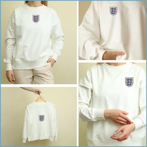 Puede incluir: Una sudadera blanca de manga larga con cuello redondo y un escudo bordado en la parte superior izquierda del pecho. El escudo muestra tres leones azules dispuestos verticalmente dentro de una forma de escudo, delineado en hilo rojo y azul.