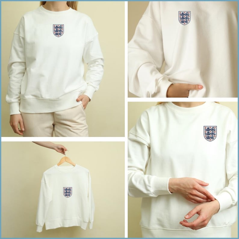 Puede incluir: Una sudadera blanca de manga larga con cuello redondo y un escudo bordado en la parte superior izquierda del pecho. El escudo muestra tres leones azules dispuestos verticalmente dentro de una forma de escudo, delineado en hilo rojo y azul.