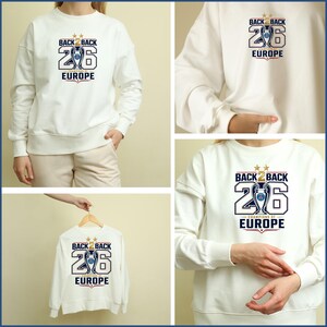 Pode incluir: Uma camisola branca de manga comprida com uma impress&atilde;o gr&aacute;fica que diz BACK 2 BACK 26 CHAMPIONS OF EUROPE, com um &iacute;cone de trof&eacute;u central, duas estrelas douradas por cima e detalhes em vermelho e azul no peito da pe&ccedil;a.