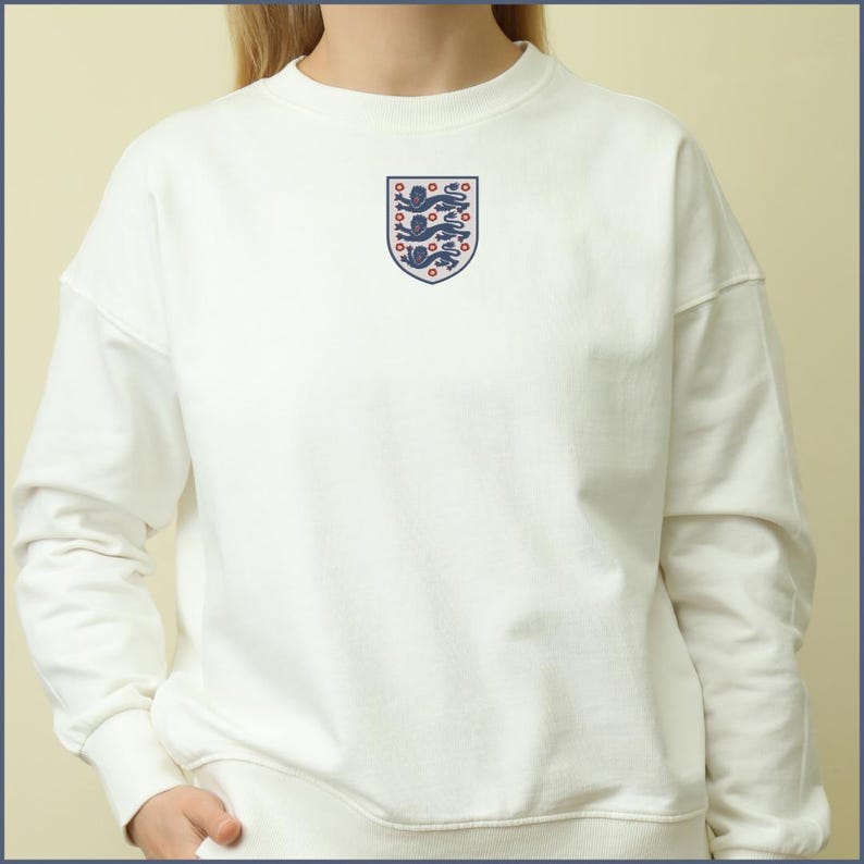 Puede incluir: Una sudadera de cuello redondo color crema con un escudo bordado en el centro. El escudo tiene forma de blas&oacute;n y contiene tres leones azules apilados con detalles en rojo, rodeados por un borde azul fino en el pecho.