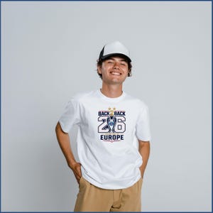 Pode incluir: Uma t-shirt branca com um gr&aacute;fico contendo o texto BACK 2 BACK, o n&uacute;mero 26 e CHAMPIONS OF EUROPE, juntamente com uma ilustra&ccedil;&atilde;o de um trof&eacute;u e duas estrelas douradas. A t-shirt &eacute; combinada com um bon&eacute; branco e preto e cal&ccedil;as bege.