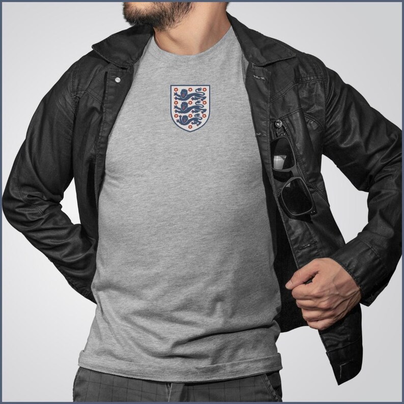 Puede incluir: Una camiseta gris jaspeado de manga corta con un emblema de escudo centrado en el pecho. El escudo muestra tres leones azules apilados con detalles rojos, delineados en azul. La camiseta se lleva debajo de una chaqueta negra con bolsillo.