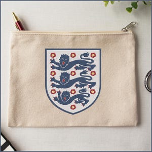 Puede incluir: Un estuche de lona beige con cremallera que presenta un emblema de escudo centrado. El escudo muestra tres leones azul oscuro en una pila vertical, rodeados por peque&ntilde;os iconos de rosas Tudor rojas y amarillas, todo en un borde azul.